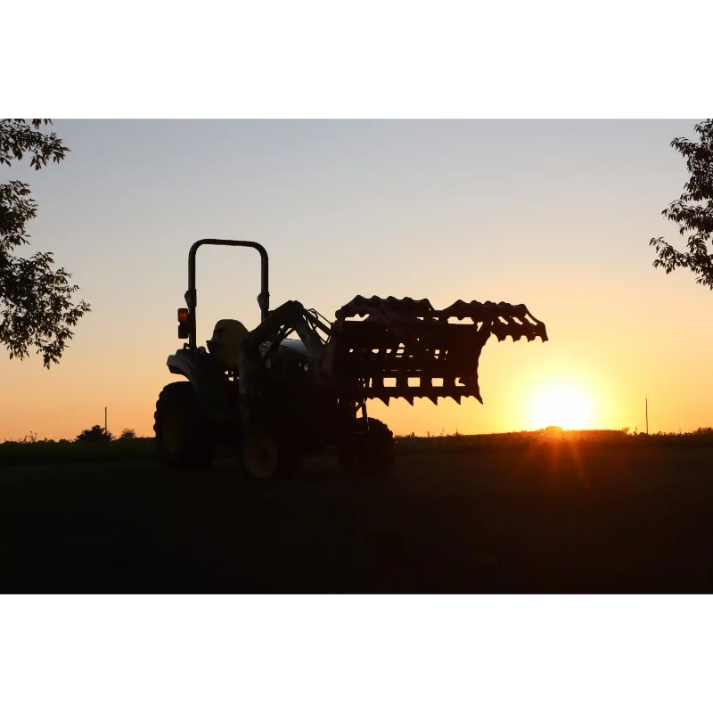 Silhouetted tractor with front loader attachment holding a large metal implement against sunset sky outdoors