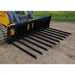 Black-painted steel manure tine bucket attachment with ten curved tines mounted on skid steer loader outdoors on dirt surface