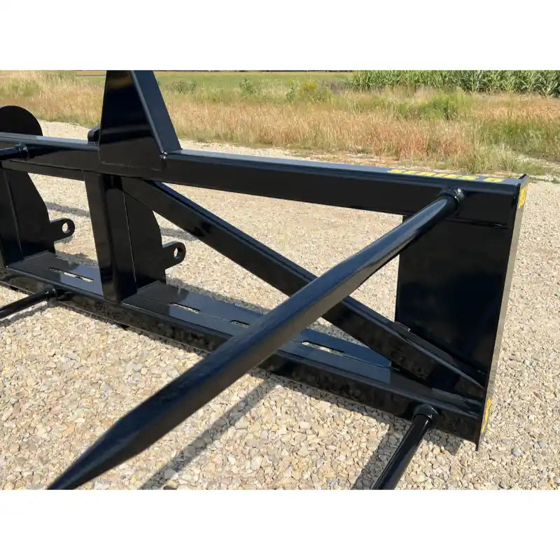 Black steel loader attachment featuring five tapered bale spears with reinforced crossbars on gravel surface outdoors