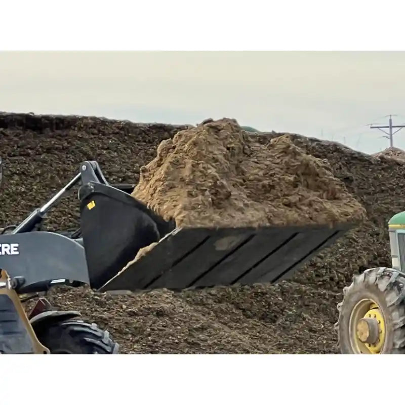 Side view of skid steer loader bucket lifting loose dirt with soil pile in background at outdoor worksite