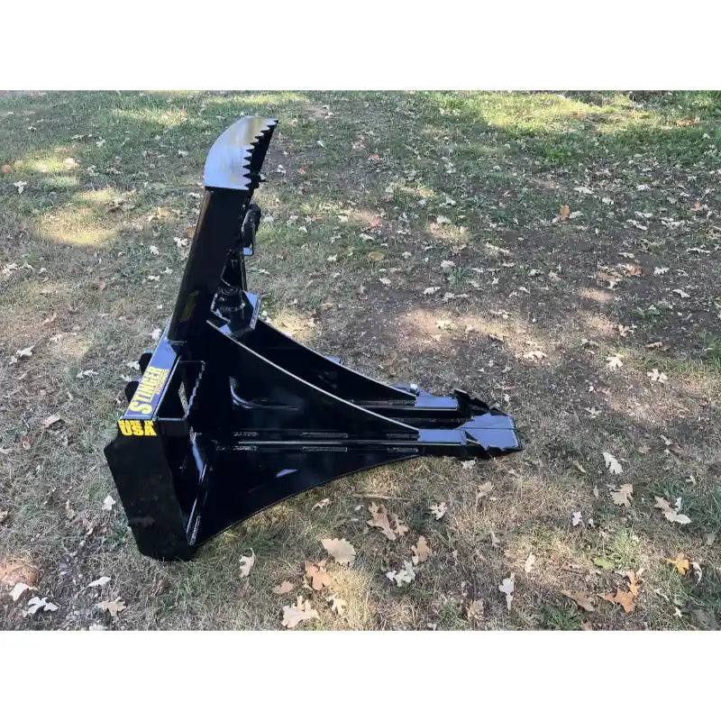 Black steel tree puller attachment with serrated jaw and triangular frame resting on grass with scattered leaves