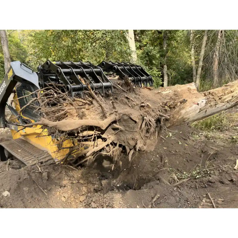 Yellow skid steer loader with black clam grapple lifting large tree stump with roots and soil outdoors