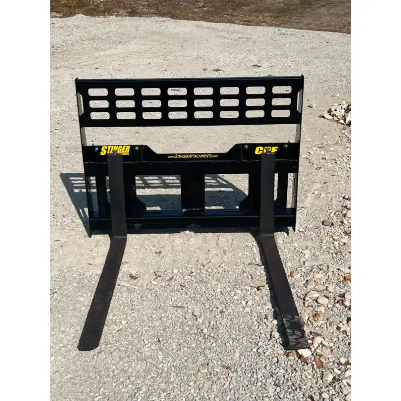 Black steel compact pallet fork attachment with perforated backrest and two extended fork tines on gravel ground outdoors