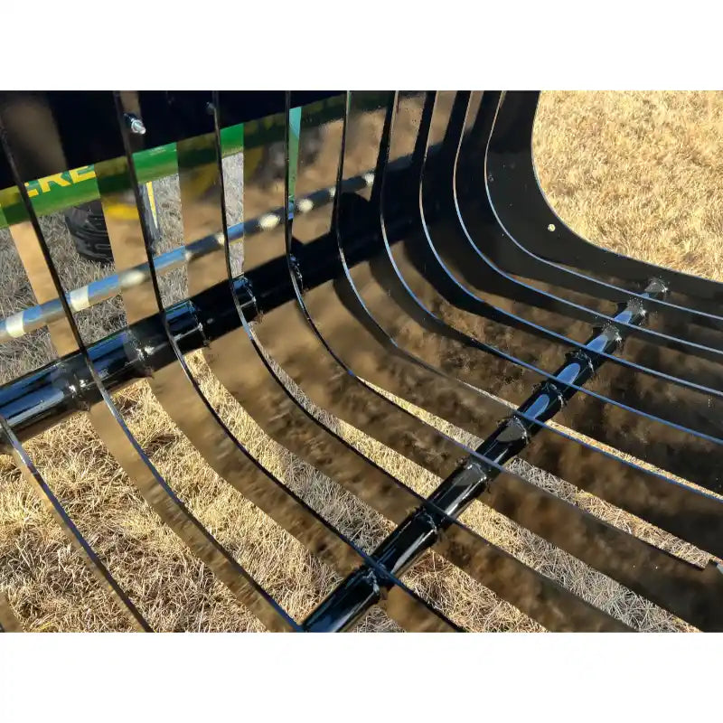 Close-up of black metal rock grapple attachment with widely spaced curved tines and reinforced crossbars over grassy ground