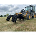 Yellow skid steer loader equipped with large black multi-tine grapple attachment on grassy field under cloudy sky