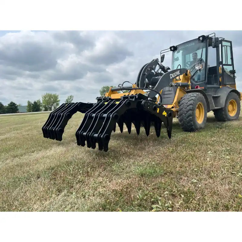 Yellow skid steer loader equipped with large black multi-tine grapple attachment on grassy field under cloudy sky