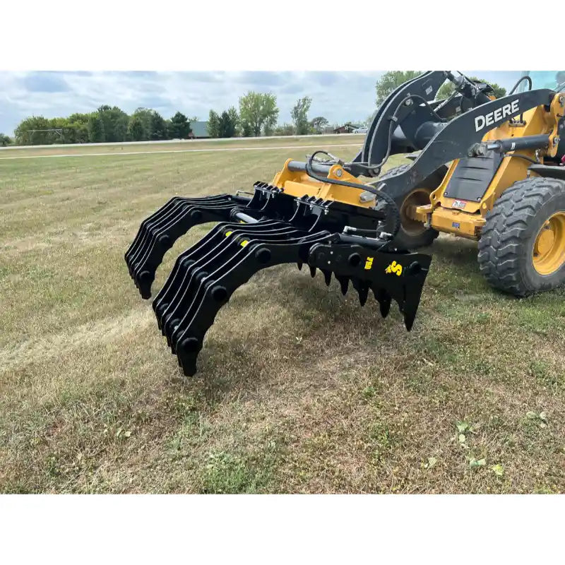 Yellow Deere skid steer equipped with black steel rake-style attachment over grassy field under cloudy sky