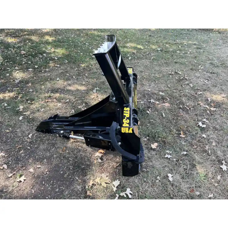 Black steel tree puller attachment with serrated gripping teeth and hydraulic arm resting on dry grass with scattered leaves