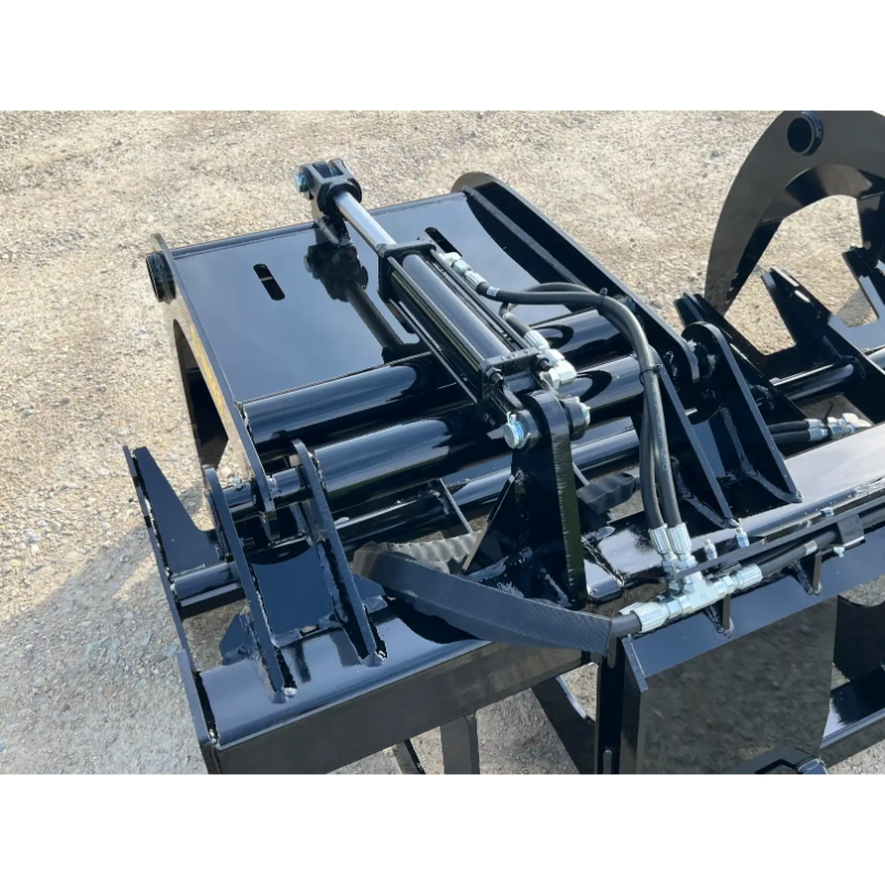Close-up of black metal skid steer hydraulic grapple attachment with hoses, cylinders, and mounting bracket on gravel surface