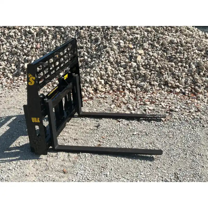 Black steel compact pallet fork attachment with welded grid backrest and two extended fork tines on gravel ground outdoors