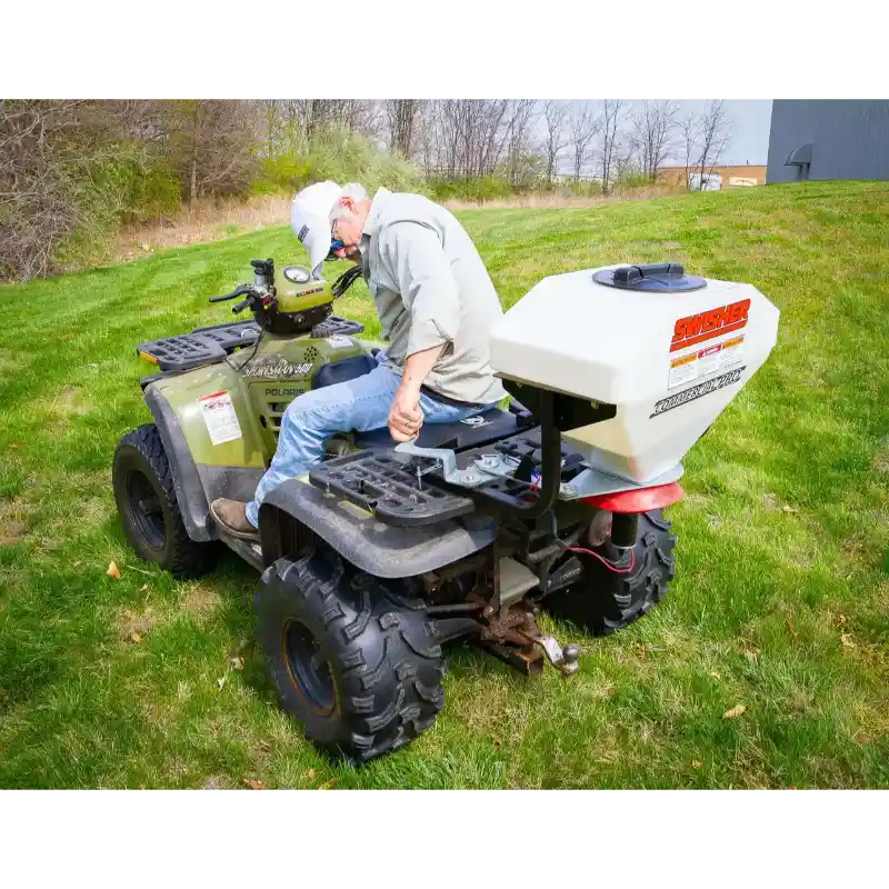 Green ATV with white Swisher spreader mounted on rear rack and operator adjusting controls, gallery position 1