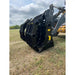 Close-up of black steel clam-style grapple attachment mounted on loader arm resting on dry grassy field under cloudy sky