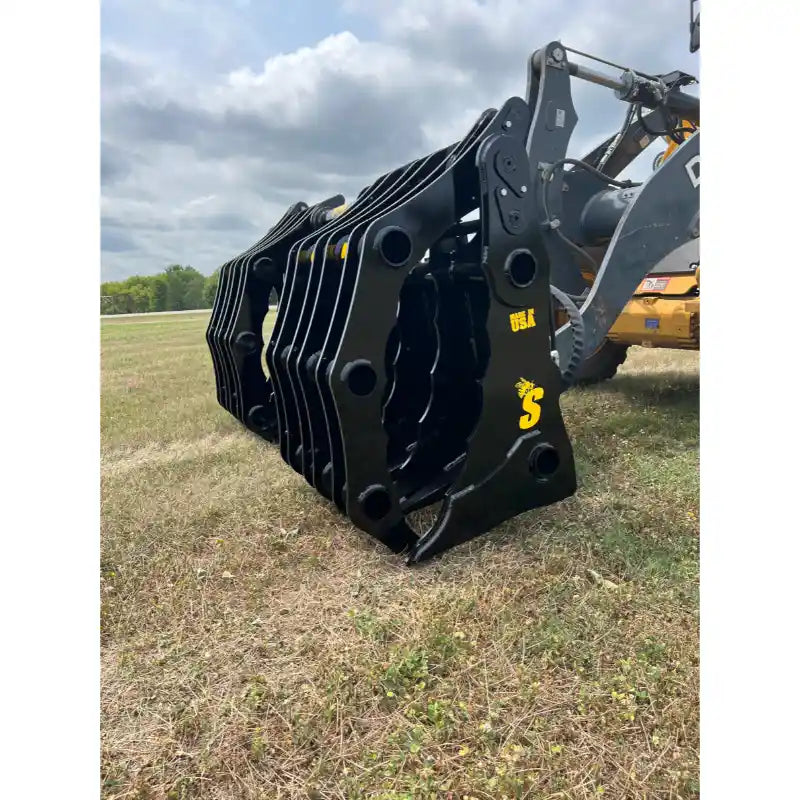 Close-up of black steel clam-style grapple attachment mounted on loader arm resting on dry grassy field under cloudy sky