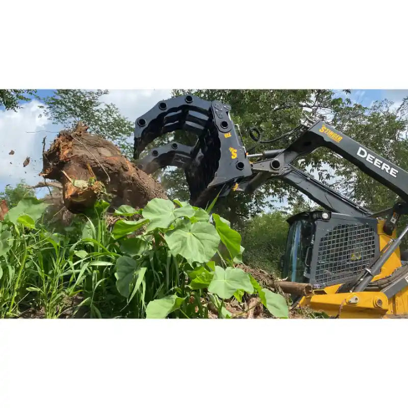 Yellow skid steer with black clam grapple attachment lifting large tree root and soil from dense vegetation