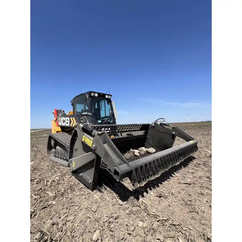 Front-right angled view of tracked skid steer with rock reel attachment on dry field under clear sky