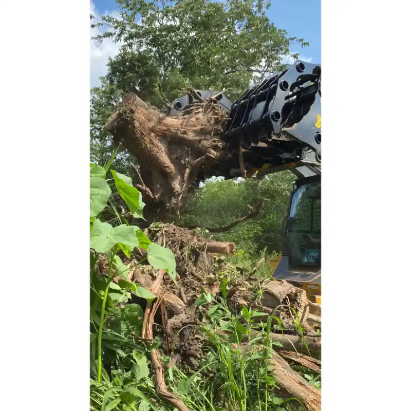 Black steel clam grapple attachment lifting large root ball with soil from overgrown vegetation near skid steer loader