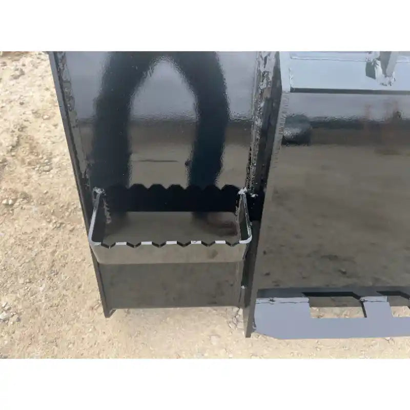Close-up of metal skid steer bucket with serrated cutting edge and step attachment on dirt surface