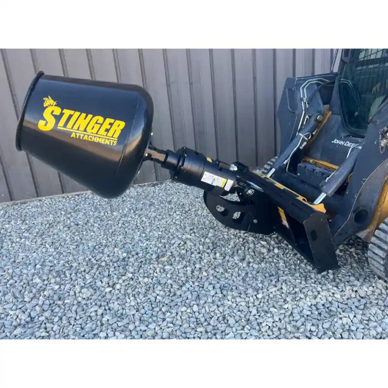 Black Stinger mixing drum attachment connected to skid steer loader arm over gravel surface near metal wall