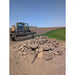 Distant view of tractor with rock bucket attachment holding pile of mixed-sized rocks on bare soil near grassy edge
