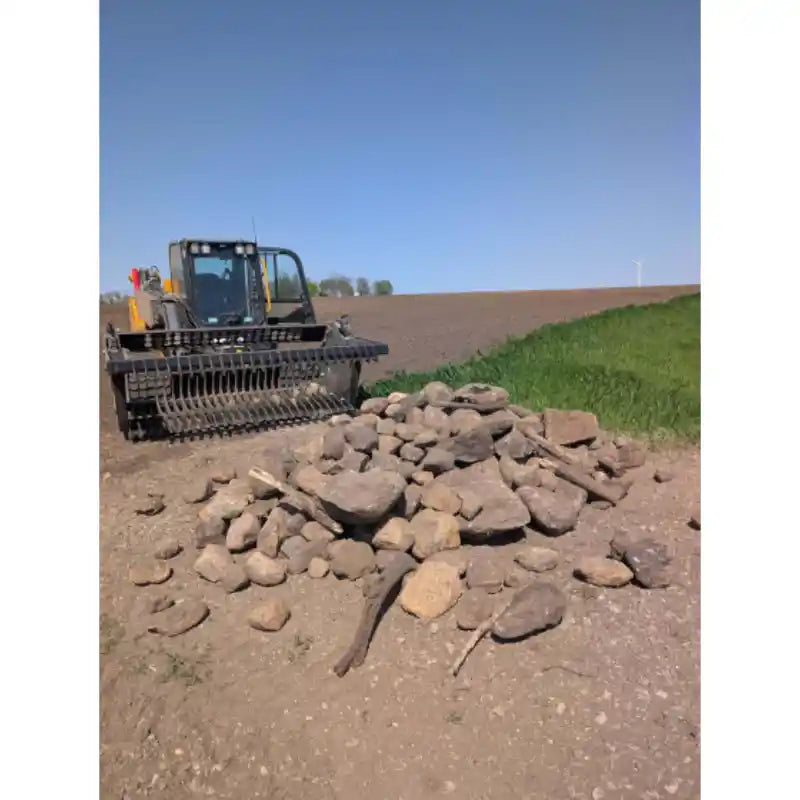 Distant view of tractor with rock bucket attachment holding pile of mixed-sized rocks on bare soil near grassy edge
