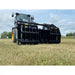 Front view of black steel clam-style grapple attachment mounted on yellow loader over grassy field under cloudy sky
