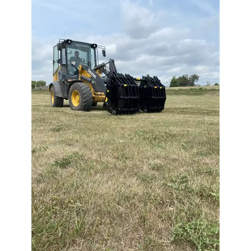 Yellow Deere loader with enclosed cab and multi-tine black grapple attachment lowered over dry grass field