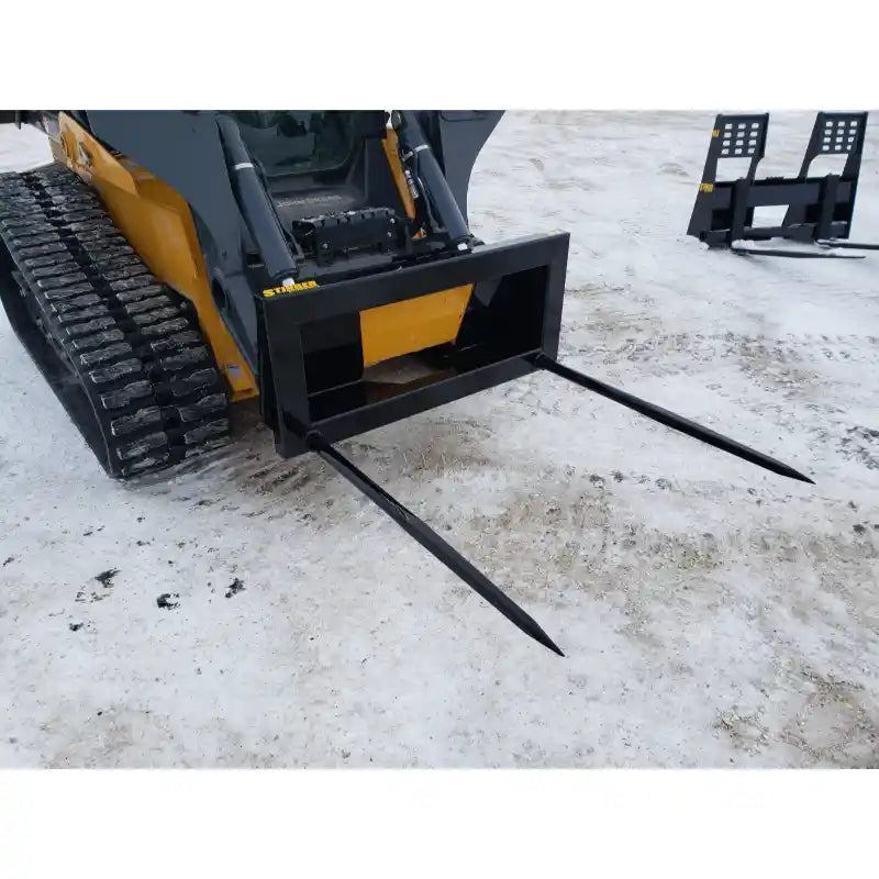 Tracked skid steer with black steel bale spear attachment featuring two long pointed tines on dirt surface