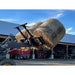 Loader attachment lifting two large round hay bales outdoors on dirt surface near farm equipment shed
