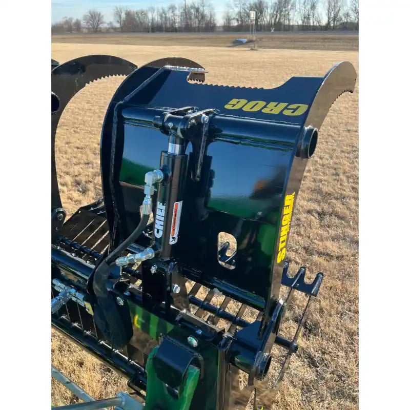 Black steel compact rock grapple attachment with serrated clamshell jaws and hydraulic hoses over dry grass