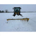 Yellow skid steer loader with wide black snow pusher blade clearing thick snow on flat open field under overcast sky
