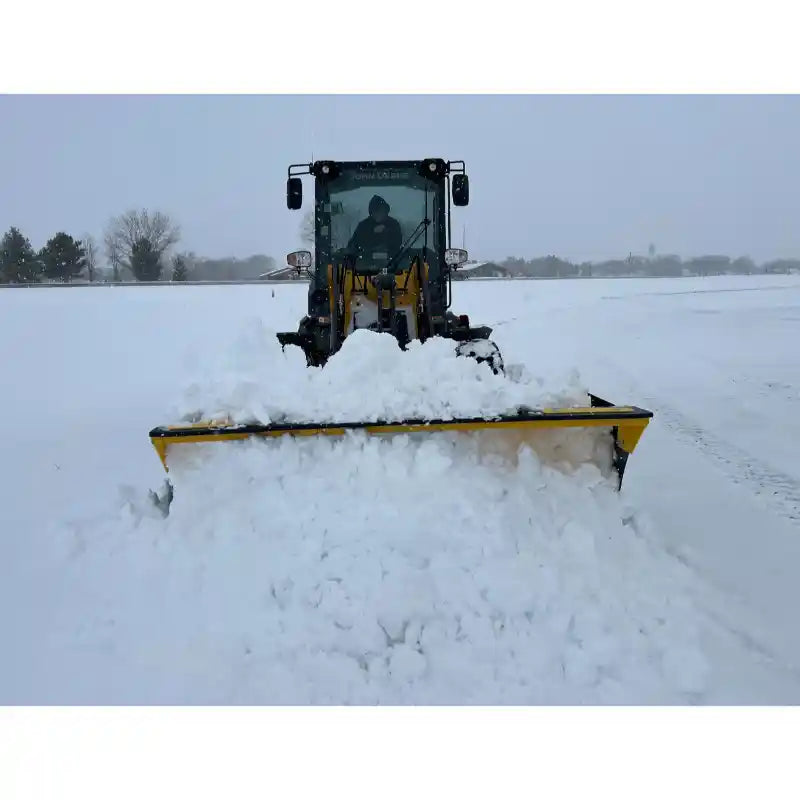 Yellow skid steer loader with wide black snow pusher blade clearing thick snow on flat open field under overcast sky