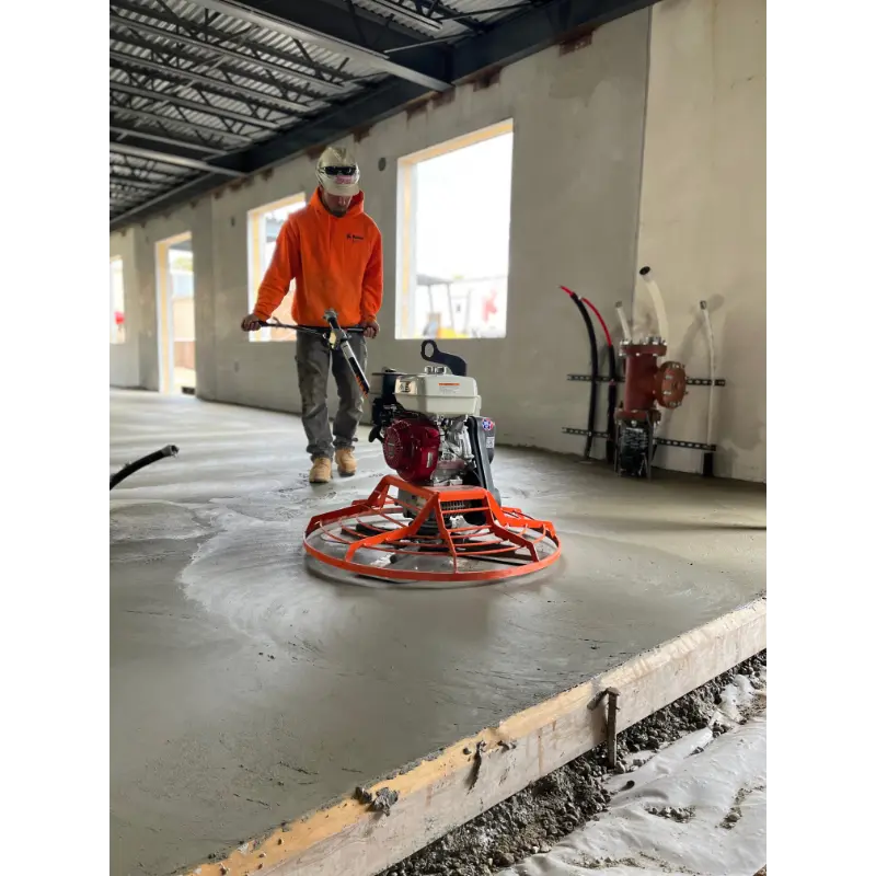 Concrete power trowel with white fuel tank and red engine operated by worker wearing orange shirt on wet concrete floor