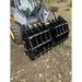 Front view of black steel clam grapple attachment mounted on skid steer loader with hydraulic hoses on dirt and grass