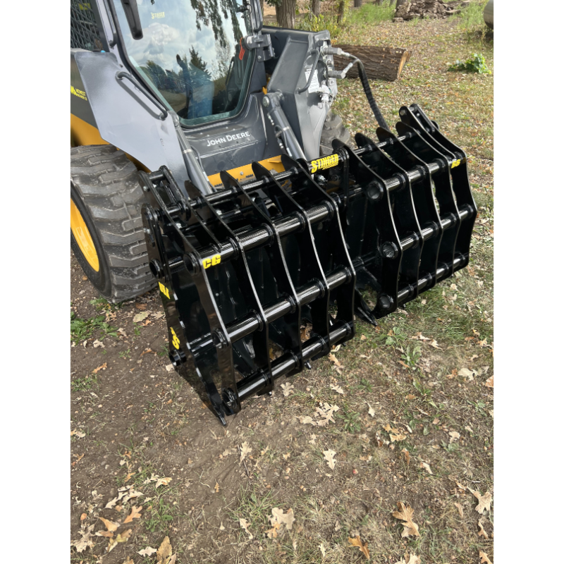 Front view of black steel clam grapple attachment mounted on skid steer loader with hydraulic hoses on dirt and grass