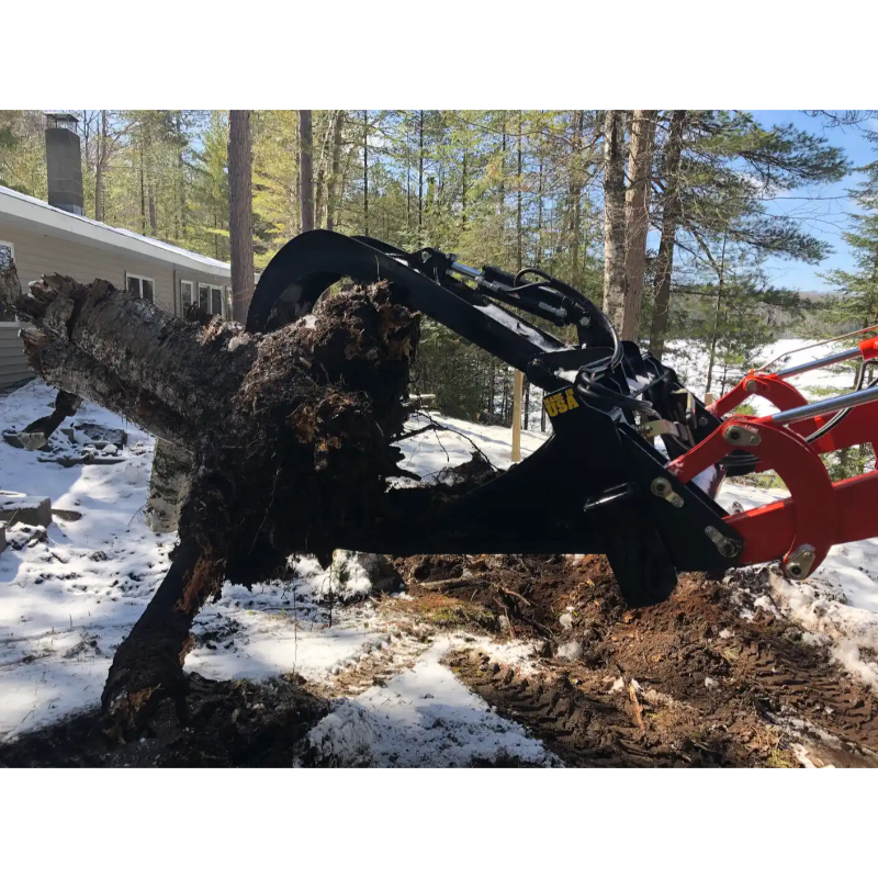 Black skid steer grapple attachment lifting large uprooted tree stump with visible hydraulic cylinders on snowy terrain