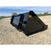Black steel skid steer material bucket with smooth sides and "Made in USA" label resting on gravel outdoors