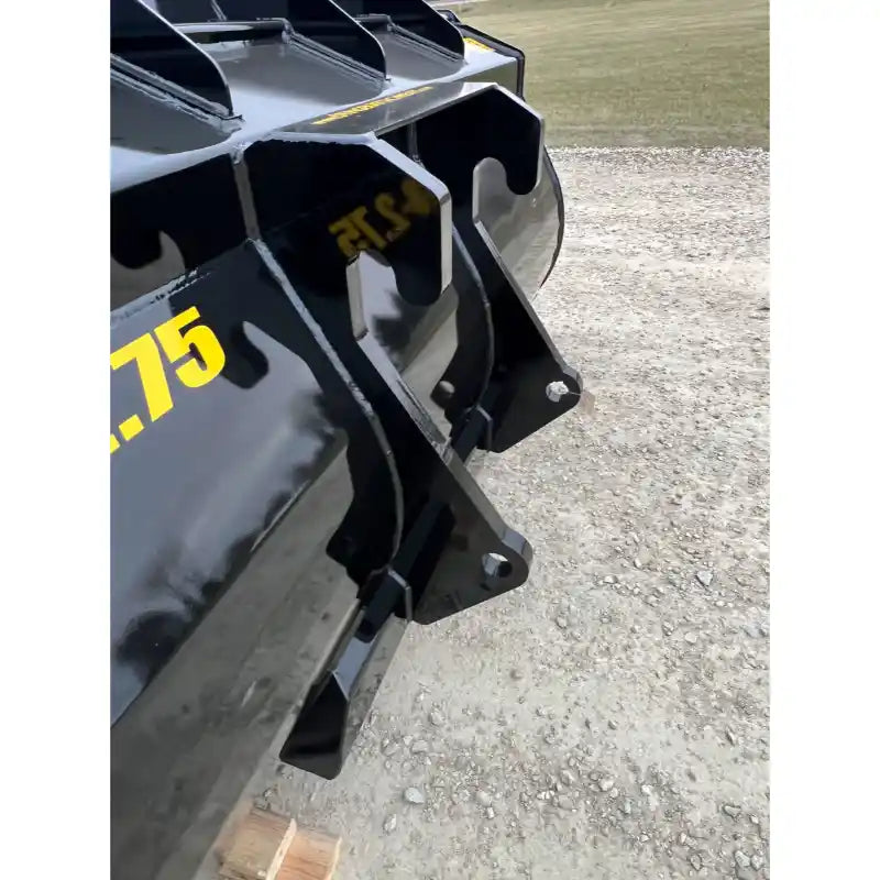 Close-up of black steel bucket attachment showing side mount brackets and part of curved bucket edge on gravel ground