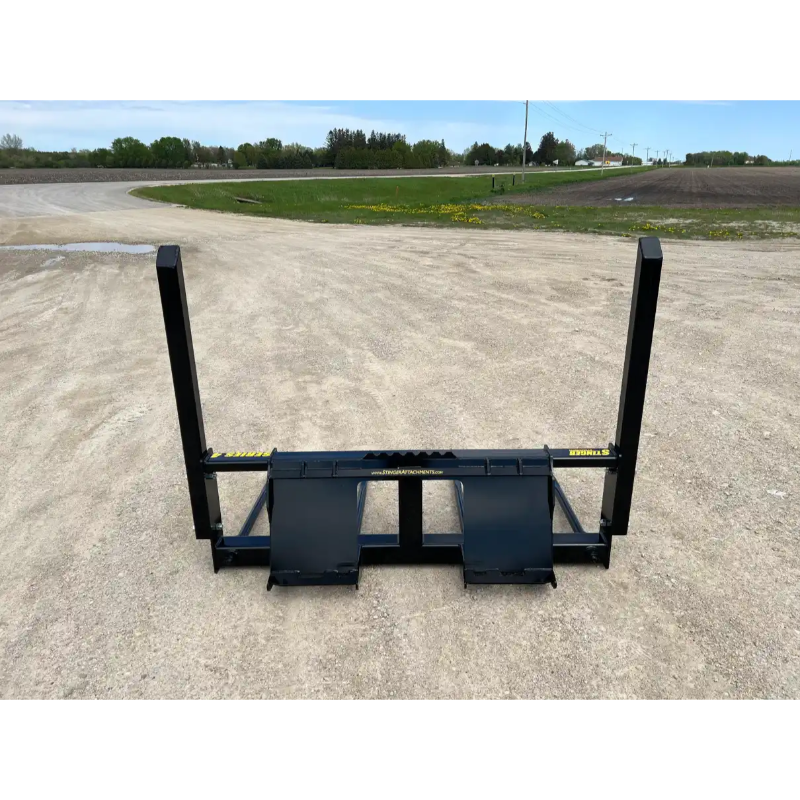 Black metal skid steer attachment with two upright arms on a gravel surface and a rural background.