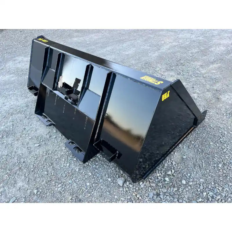 Black steel tractor attachment bucket with reinforced back and mounting brackets resting on gravel surface outdoors