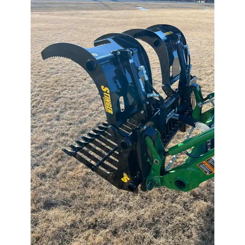 Black steel rock grapple attachment with serrated curved jaws and spaced flat tines mounted on green loader arm over dry grass field