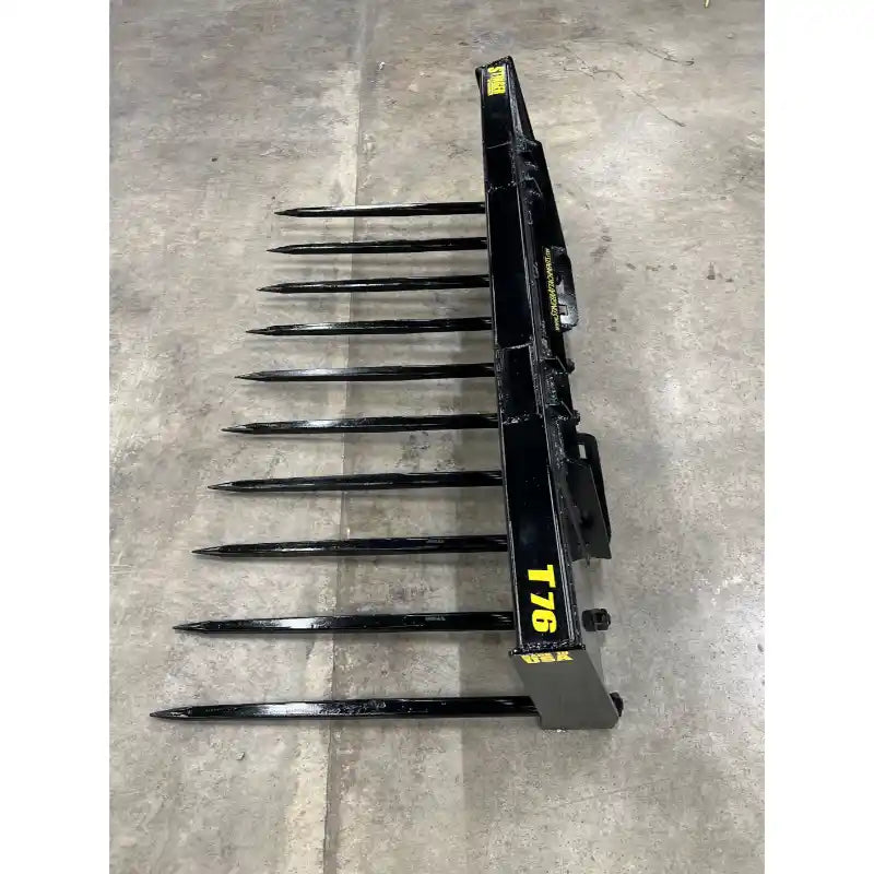 Top view of black steel manure spear attachment with ten long pointed tines on concrete floor indoors