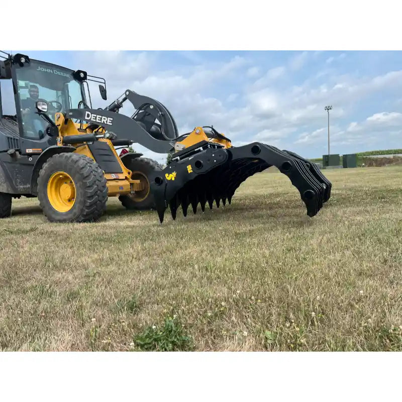 Yellow John Deere loader fitted with black steel grapple attachment lowered over dry grass field under cloudy sky