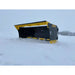 Black and yellow skid steer snow pusher attachment resting on snow-covered terrain with overcast sky background