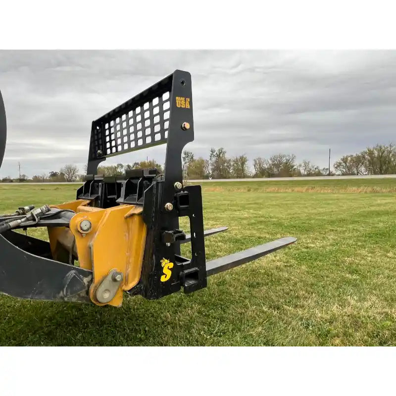Yellow skid steer loader with black steel pallet fork attachment and perforated vertical backrest on grassy field
