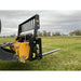 Yellow skid steer loader with black steel pallet fork attachment and perforated vertical backrest on grassy field
