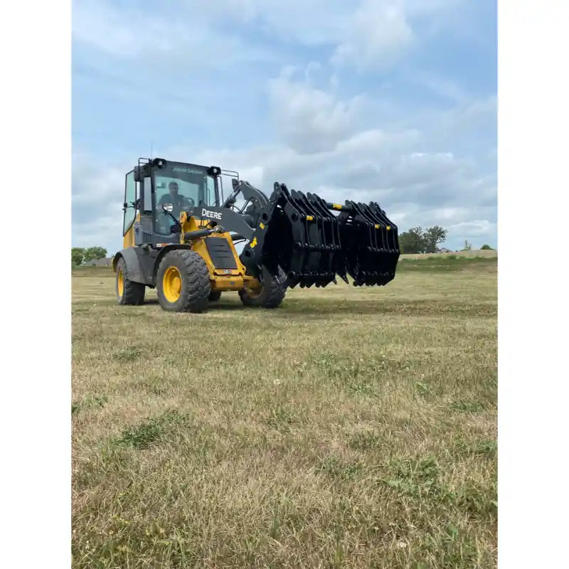 Yellow John Deere loader lifting large black clam-style grapple attachment over grassy field under partly cloudy sky