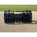 Black steel grapple attachment with multiple curved tines and central bucket resting on gravel outdoors