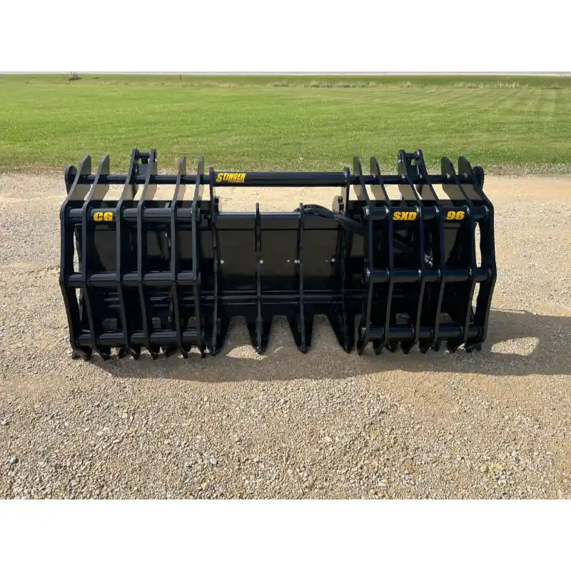 Black steel grapple attachment with multiple curved tines and central bucket resting on gravel outdoors