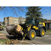 Yellow Komatsu loader with black clam-style grapple attachment lifting large tree log on paved street near buildings