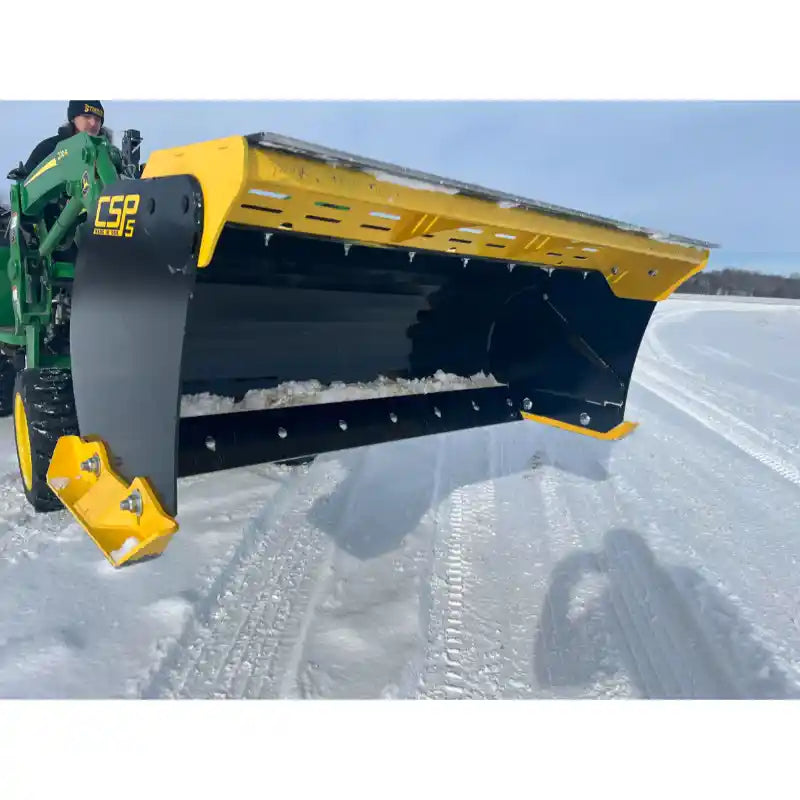 Yellow and black steel bucket attachment with cutting edge mounted on green compact track loader over snow-covered terrain